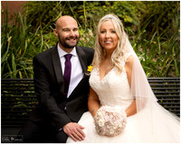 A bride and groom smile at the camera seated on a bench in front of greenery in Windsor's smallest public park