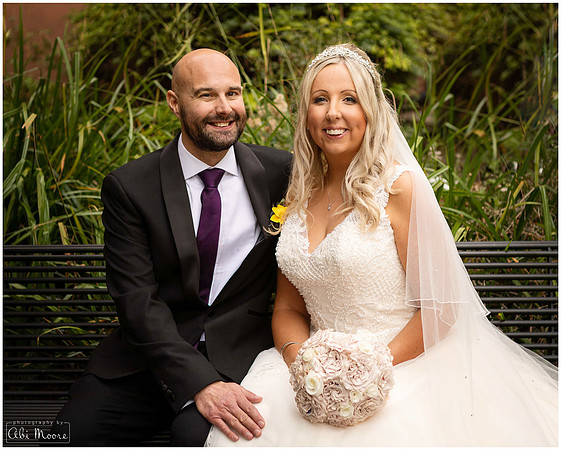 A bride and groom smile at the camera seated on a bench in front of greenery in Windsor's smallest public park