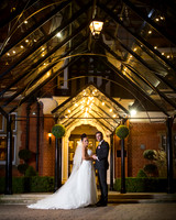 A wedding couple smile whilst standing in the lit glass porch of the Berystede at night time