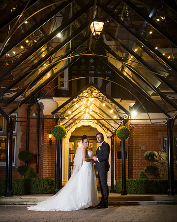 A wedding couple smile whilst standing in the lit glass porch of the Berystede at night time