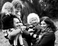 A family of five laugh together, the father holds two children on his shoulders whilst mum carries a third, photographed in black & white