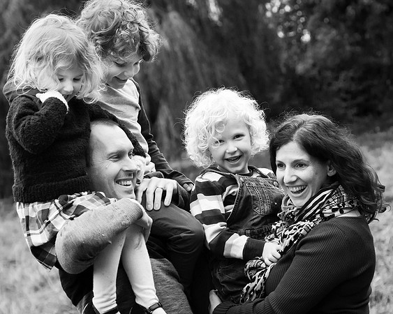 A family of five laugh together, the father holds two children on his shoulders whilst mum carries a third, photographed in black & white