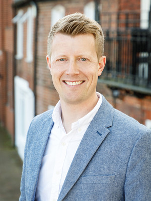 A professional headshot taken outdoors near offices in Windsor Berkshire, a businessman smiles at the camera wearing a blue suit with old victorian office buildings behind.