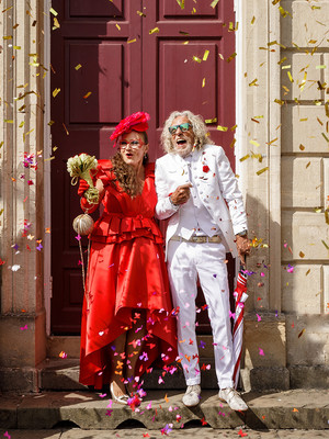 Bride & groom smile and laugh outside the red doors of the Windsor Guildhall as confetti falls around them, the couple are in later life, the bride is in red and the groom is in white