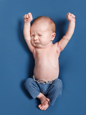 A newborn baby dressed in blue trousers and no top stretches sleepily, laying on a blue backdrop that matches his trousers