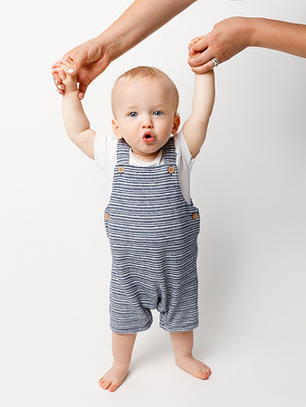 A baby aged around one year stands whilst holding his parent's hands (just in shot) and makes a cue 'ooh' expression