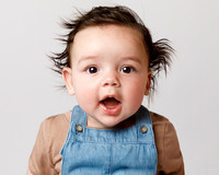 A baby with lots of crazy black hair sticking out is making a cute expression, photographed in close-up head and shoulders