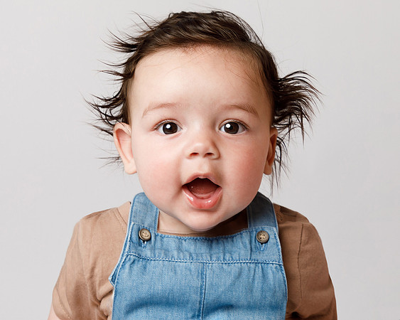 A baby with lots of crazy black hair sticking out is making a cute expression, photographed in close-up head and shoulders