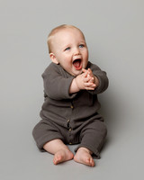A baby aged about nine months sits in studio photography setting and claps his hands whilst looking up and to the right