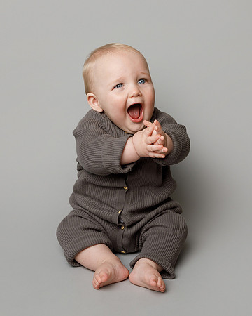 A baby aged about nine months sits in studio photography setting and claps his hands whilst looking up and to the right