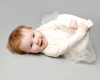 A baby girl lays on her back, holding her toes, looking straight at the camera and smiling