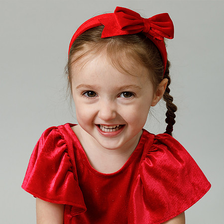 A three-year-old girl gives a big smile to the camera, dressed in red with a red bow on her hair