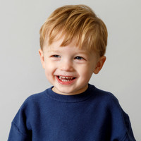 A toddler boy smiles looking just off-camera in a blue jumper on a grey studio backdrop