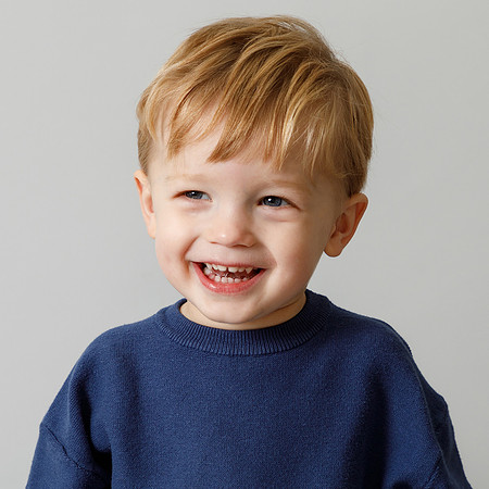 A toddler boy smiles looking just off-camera in a blue jumper on a grey studio backdrop