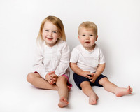 Two siblings - a four-year-old girl and her toddler brother, photographed on a white studio backdrop, dressed in white, sat next to each other and smiling
