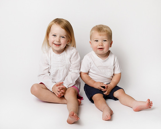 Two siblings - a four-year-old girl and her toddler brother, photographed on a white studio backdrop, dressed in white, sat next to each other and smiling