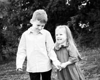 A brother and sister photographed outdoors in black & white, holding hands and smiling widely