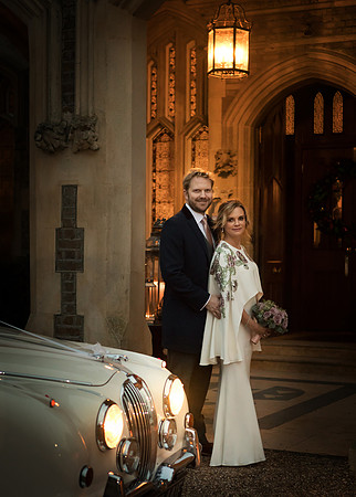 A wedding couple stand in the doorway of Oakley Court by a classic car at night time