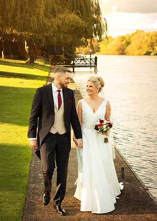 A bride and groom stroll along the riverside at Oakley Court