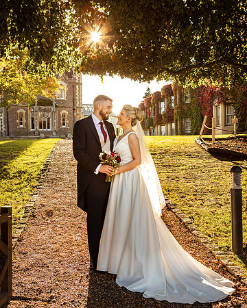 A bride and groom smiles at each other as the autumn sun shines through the trees in the grounds of Oakley Court