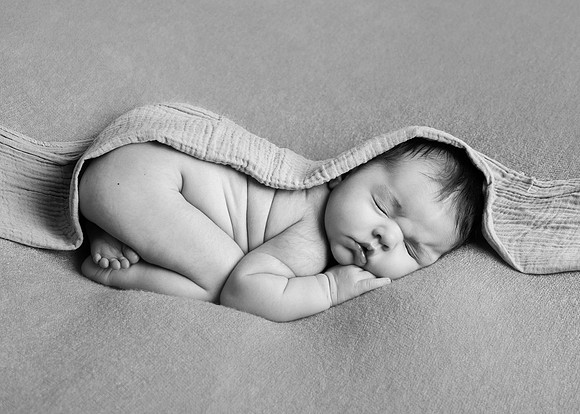 Newborn baby asleep curled up on her front with a blanket draped over her photographed in black & white