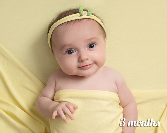 Three month old baby girl laid on yellow smiles straight at the camera