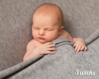 Three week old baby lays on his back asleep tucked under a grey blanket