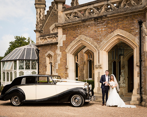 A wedding couple stand outside the gothic front of Oakley Court in Windsor, alongside a classic car