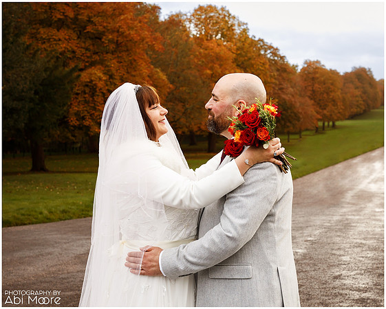 A wedding couple smile at each other hands on each others hips and shoulders, photographed close-up with autumnal trees on Windsor's Long Walk in the background