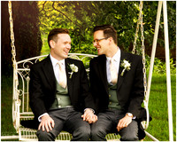 Two grooms sit on a garden swing together holding hands on their wedding day