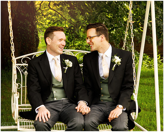 Two grooms sit on a garden swing together holding hands on their wedding day