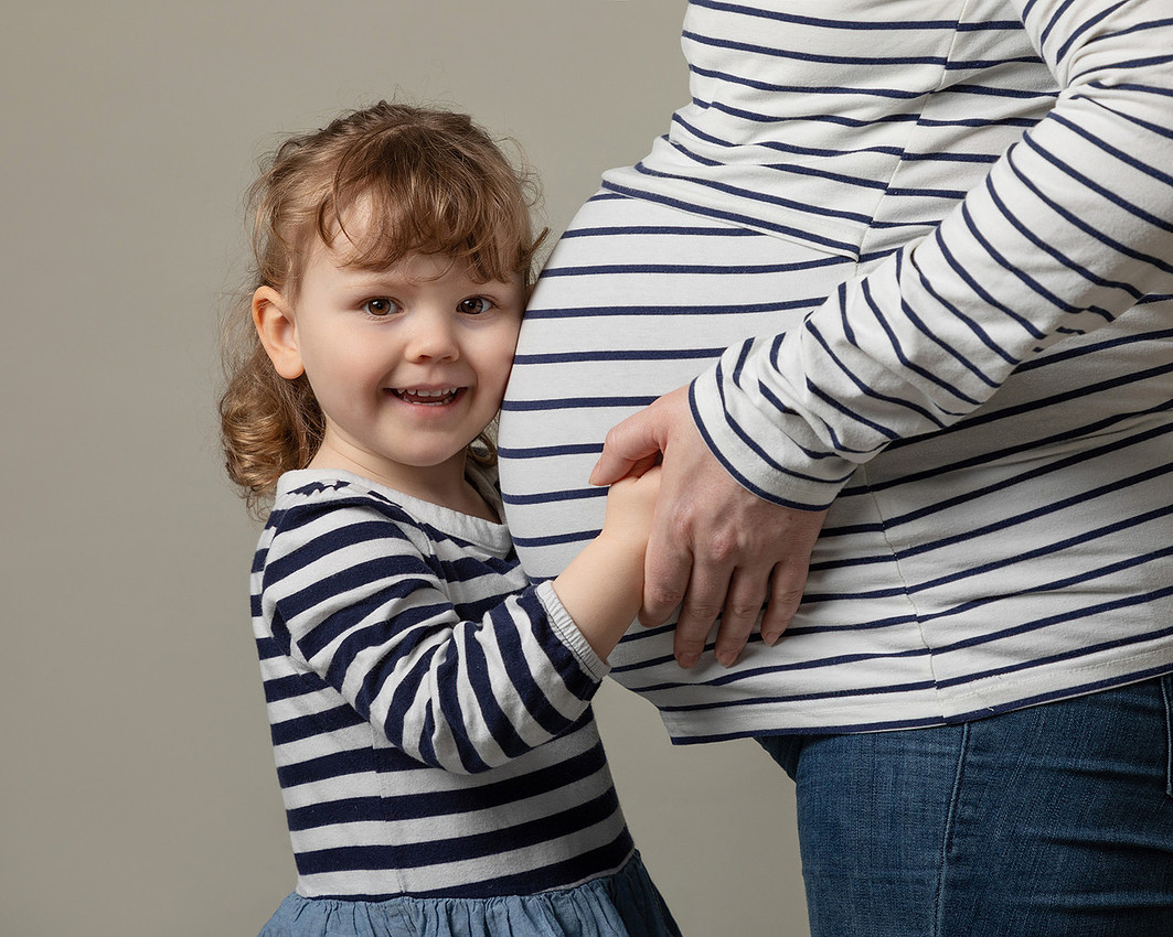 Close-up of pregnant woman's baby bump with a toddler girl pressing her ear to the bump and smiling at tehe camera, photographed in a studio setting.