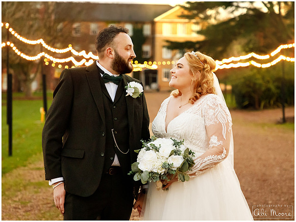 A bride and groom smile at each other lit by outdoor fairy lights all around them