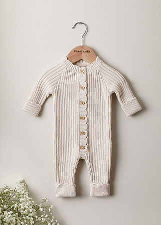 a cream knitted baby romper hangs on a wooden hanger against a neutral wall with small white flowers in the corner of the image