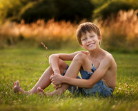 Young boy smiles in a field holding a blade of grass, reminicent of Huck Finn