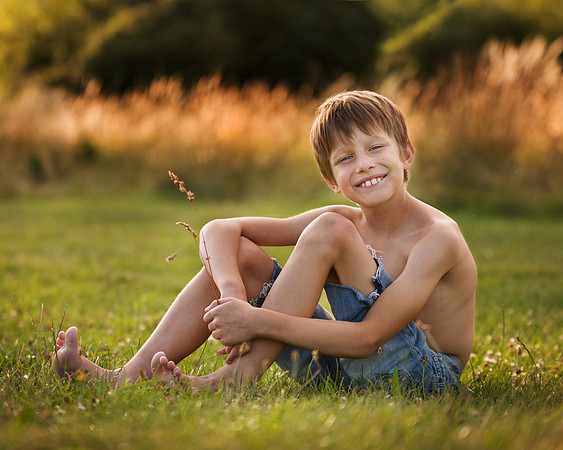 Young boy smiles in a field holding a blade of grass, reminicent of Huck Finn