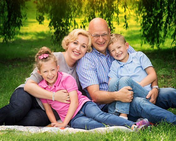 Family sit on picnic blanket and hug, smiling at camera, taken underneath a willow tree in the summer