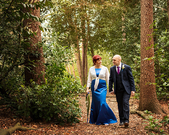 A bride and groom smile at each other whilst strolling through woodland at the Berystede in Ascot; the bride wears a stunning royal blue dress