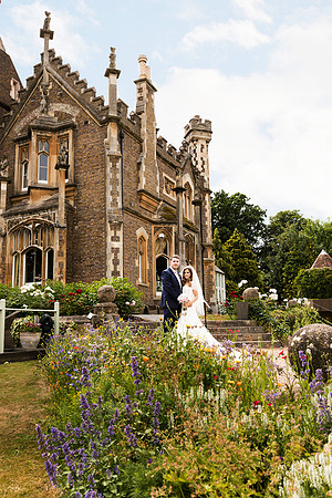 A wedding couple stand in front of gothic Oakley Court surrounded by wildflowers