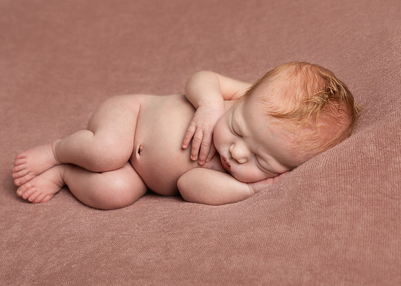 Newborn baby girl lays on her side sleeping serenely on a pink blanket