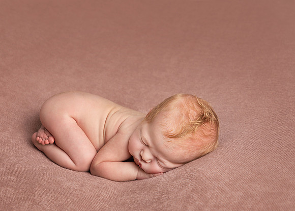 Newborn baby girl lays curled up on her front, filling only a quarter of the image, emphasising how tiny she is