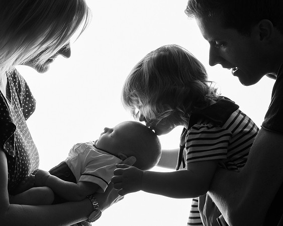 Family shot in profile and in backlit semi-silhouette - a toddler sister kisses her newborn brother on the head