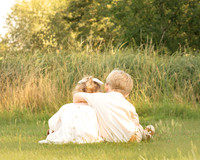 A 3-year-old boy and girl hug in a long grass meadow with their backs to the camera