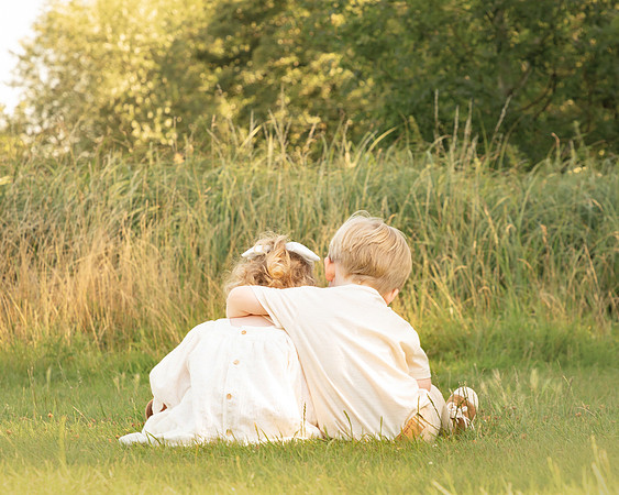 A 3-year-old boy and girl hug in a long grass meadow with their backs to the camera
