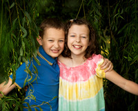 A boy and girl smile as they push through the hanging branches of a weeping willow