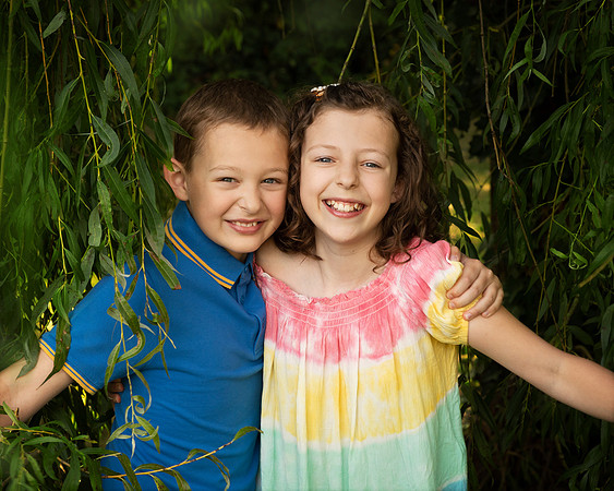 A boy and girl smile as they push through the hanging branches of a weeping willow
