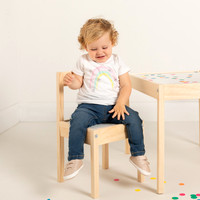 Toddler girl modelling jeans and t-shirt looks down and smiles whilst sat on a chair at a small table surrounded by confetti