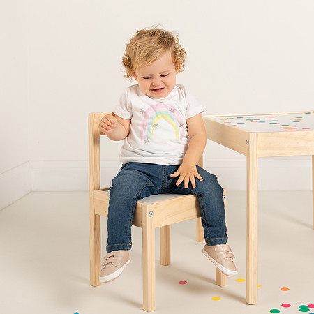 Toddler girl modelling jeans and t-shirt looks down and smiles whilst sat on a chair at a small table surrounded by confetti