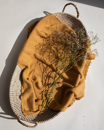 A honey-coloured blanket laid in a basket with baby's breath flowers laid on top - the bright sunlight creates defined black shadows