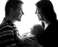 A black & white backlit semi-silhouette photo of parents with their newborn baby